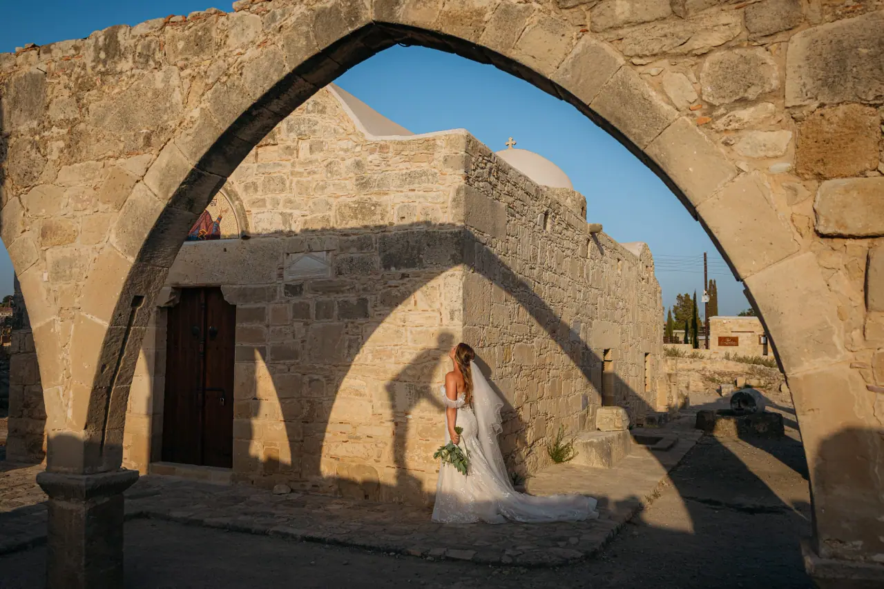 Bride framed by a crumbling stone arch beside a small stone chapel under blue sky