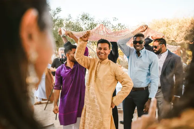 Groom's procession led by men in purple and gold kurtas beneath the canopy, Thalassines Beach Villas
