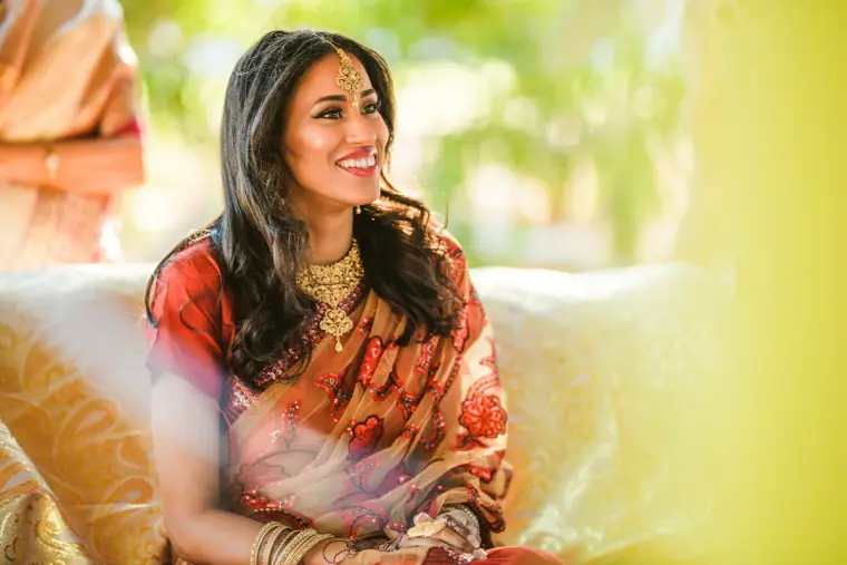 Raya in her red sari and gold jewellery smiling as she watches the groom arrive