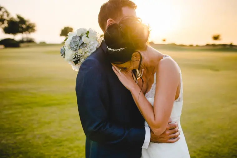 Couple embracing on a golf course lawn at sunset, bouquet raised, Aphrodite Hills