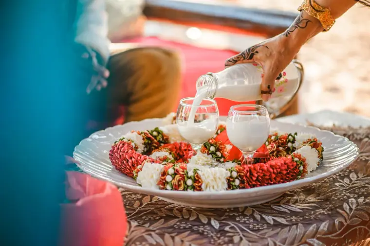 Hennaed hand pouring milk into glasses over a plate of flower garlands