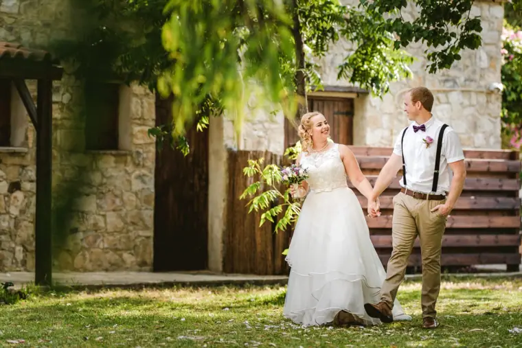 Bride and groom in braces walking hand in hand across a lawn by stone buildings