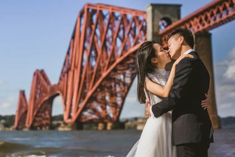 Couple kissing on the shore in front of a vast red steel railway bridge in Scotland