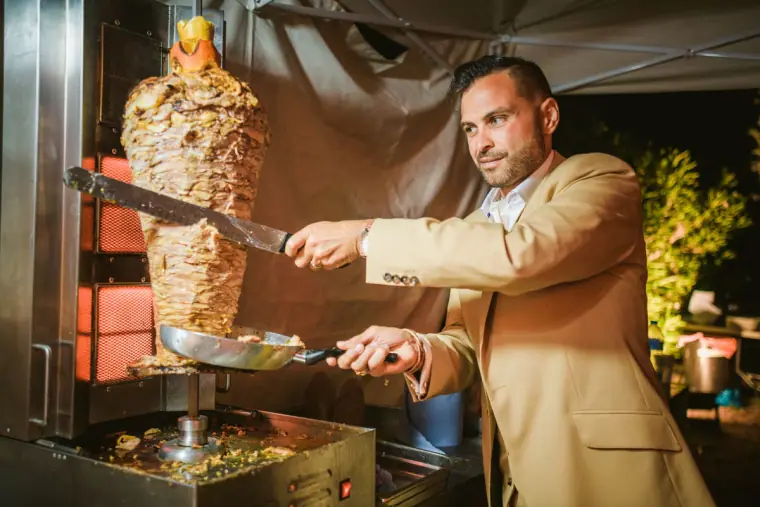 Groom in a tan jacket carving a gyros spit at an evening food station