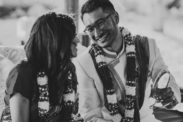 Black-and-white photo of the couple in flower garlands smiling at a hand mirror