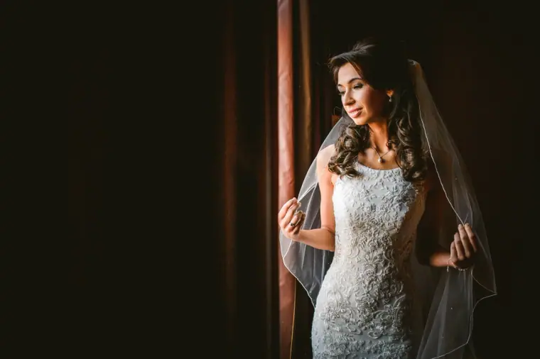 Bride in her lace gown and veil standing in soft window light