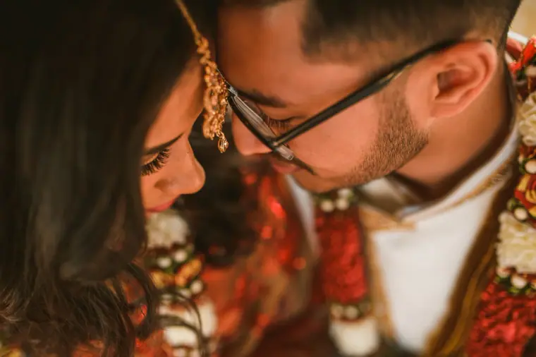 Bride and groom leaning their heads together, wearing red and white garlands