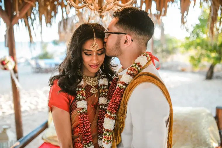 Groom kissing the bride's forehead under the thatched canopy, Thalassines Beach Villas