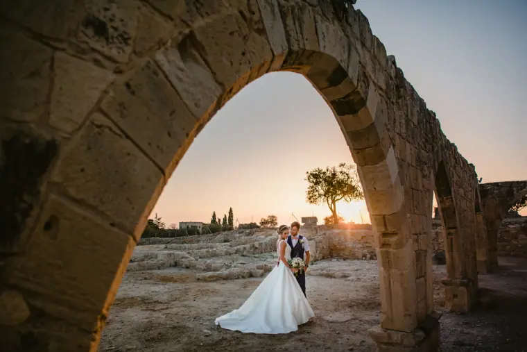 Bride and groom framed by an ancient stone arch at sunset
