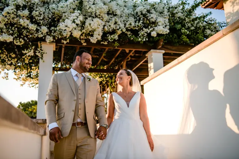 Groom in a beige suit holding hands with his bride under a white blossom pergola