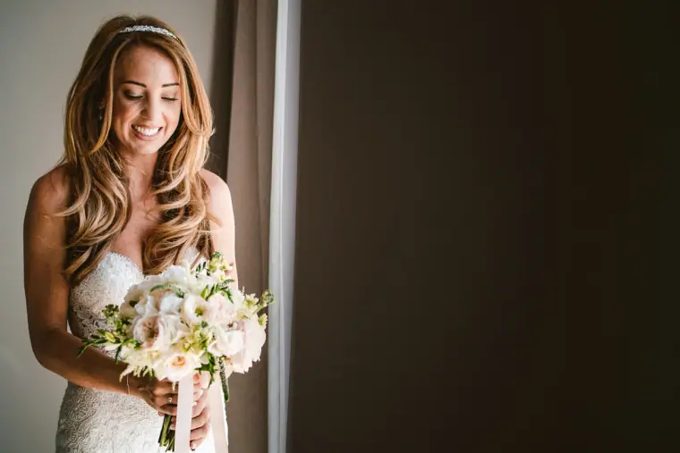 Smiling bride with long wavy hair holding a white bouquet by a window
