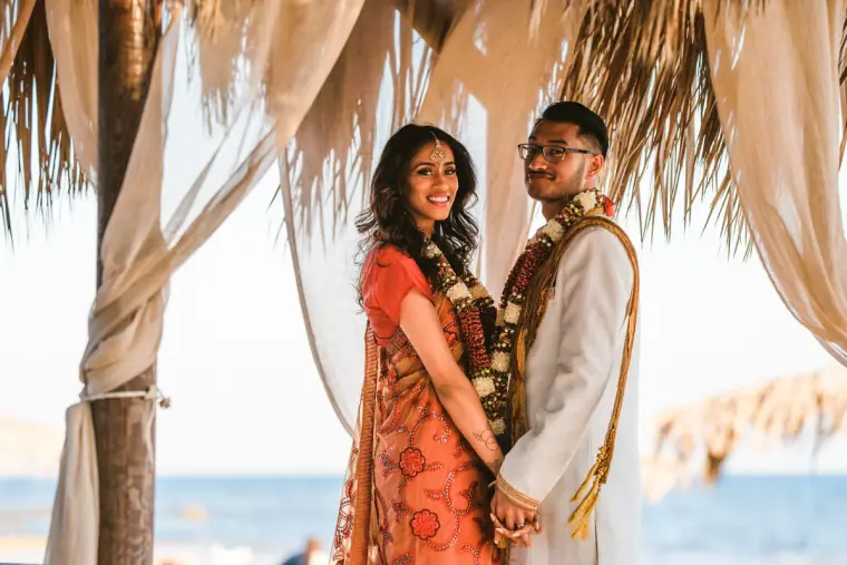 Couple in garlands standing hand in hand beneath a thatched cabana by the sea
