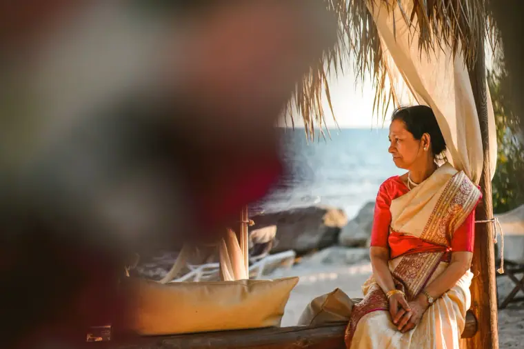 Older woman in a cream and red sari sitting on a beach daybed, looking out to sea