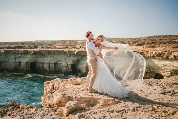 Groom lifting his bride on a sea-cave clifftop as her veil streams in the wind