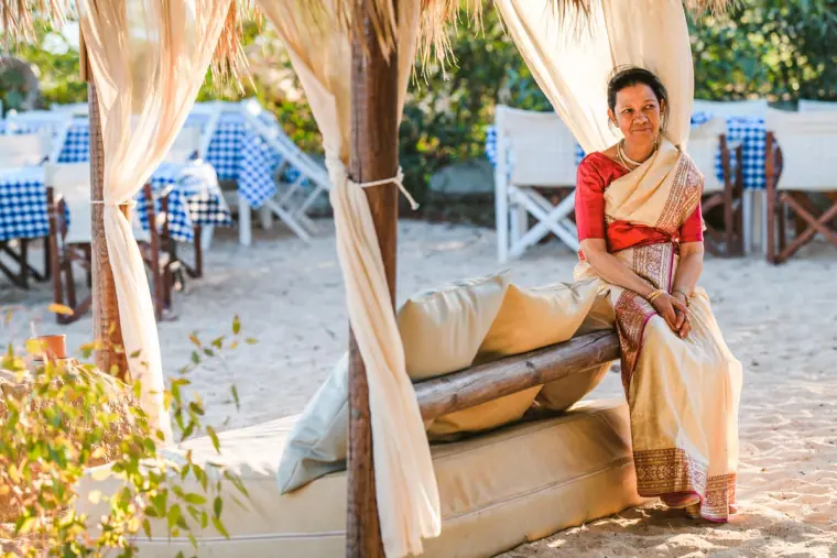 Woman in a red and cream sari seated on a curtained daybed on the sand