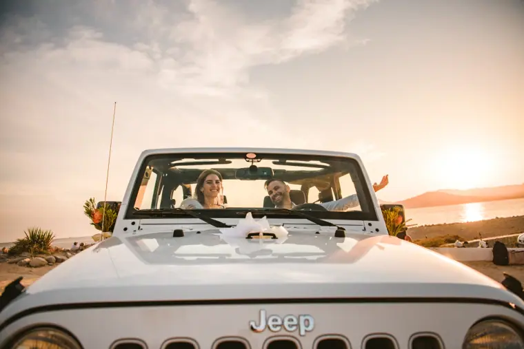 Couple in a white Jeep with its top down, sun setting over the sea behind