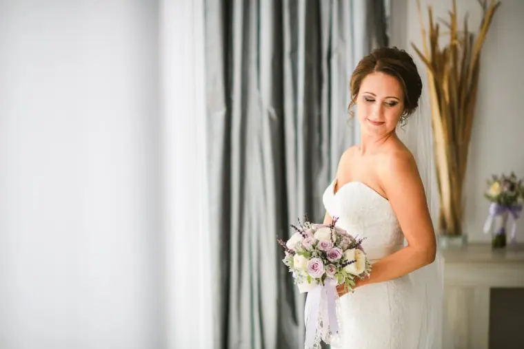 Bride holding a lilac bouquet by a bright window with grey curtains, Paphos