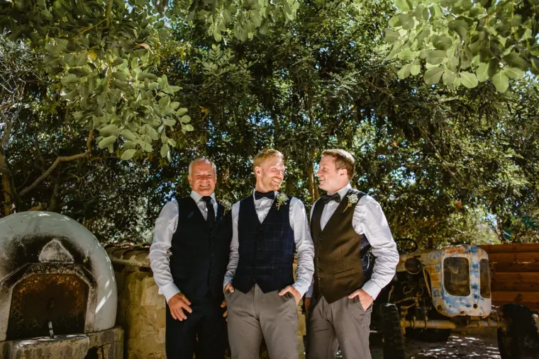 Three groomsmen in waistcoats and bow ties standing under trees