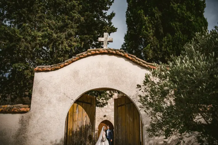 Couple kissing inside the arched gateway of a whitewashed chapel among cypress trees