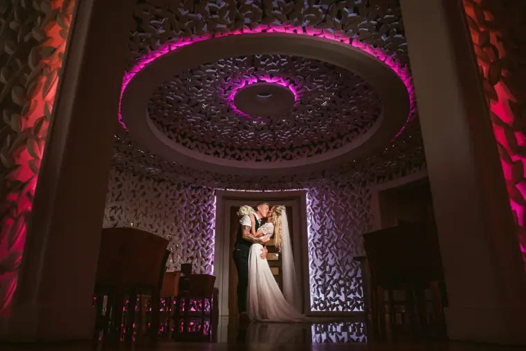 Couple embracing in a doorway beneath an ornate ceiling lit with magenta light