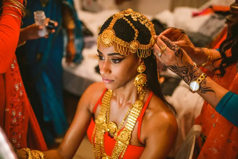 Bride in an ornate gold headdress and jewellery being dressed by relatives