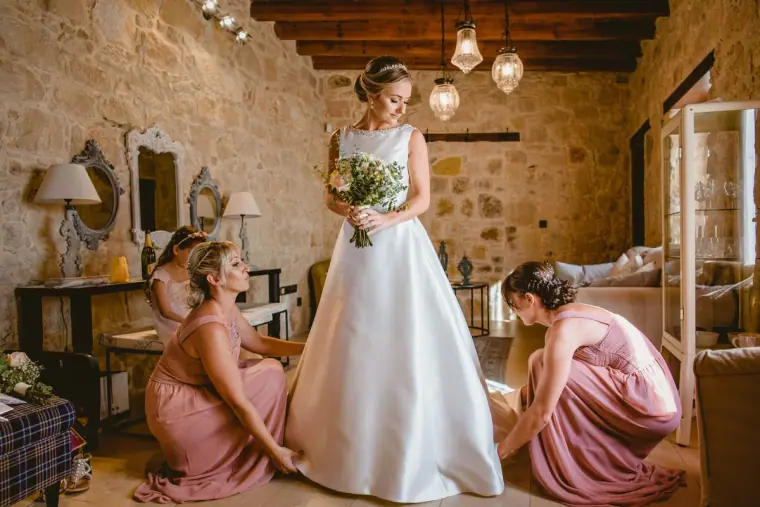 Bridesmaids in blush pink adjusting the bride's satin gown in a stone-walled room
