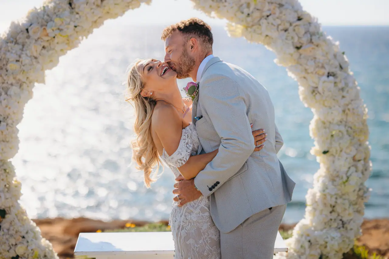 Nick kissing Jayley beneath a white floral hoop with the sea glittering behind
