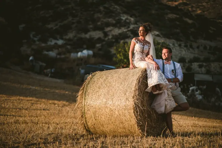 Bride perched on a round hay bale in a golden field with the groom beside her