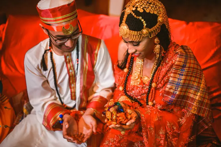 Groom in a red cap and bride in a gold headdress holding hands over a tartan shawl