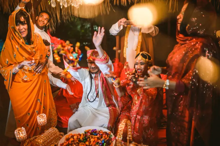 Women in orange and red cheering around the seated couple during the evening celebrations