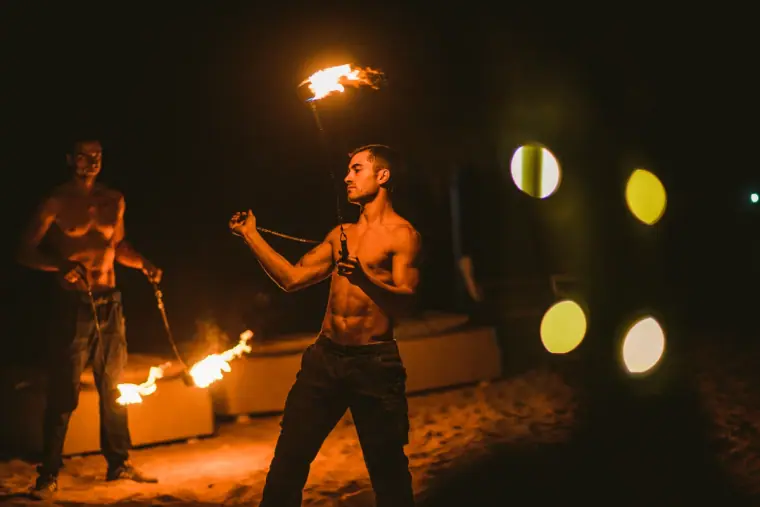 Two fire dancers performing with flaming poi on the sand at night