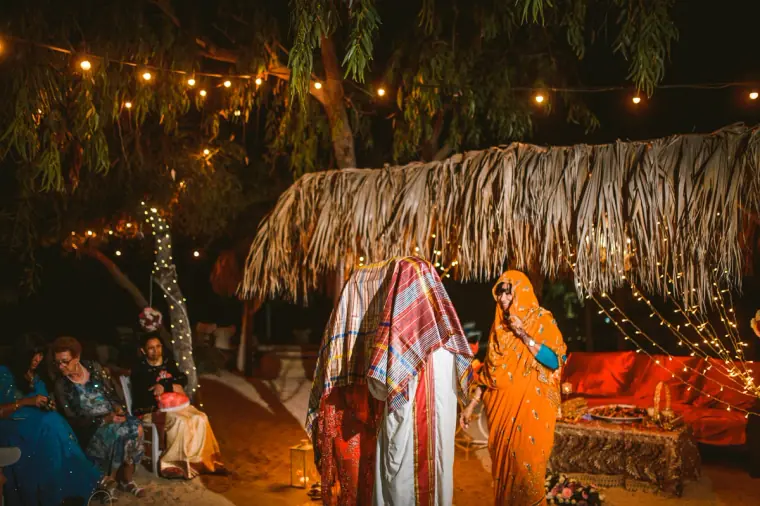 Bride wrapped in a tartan shawl beside a woman in orange beneath the lit thatched canopy
