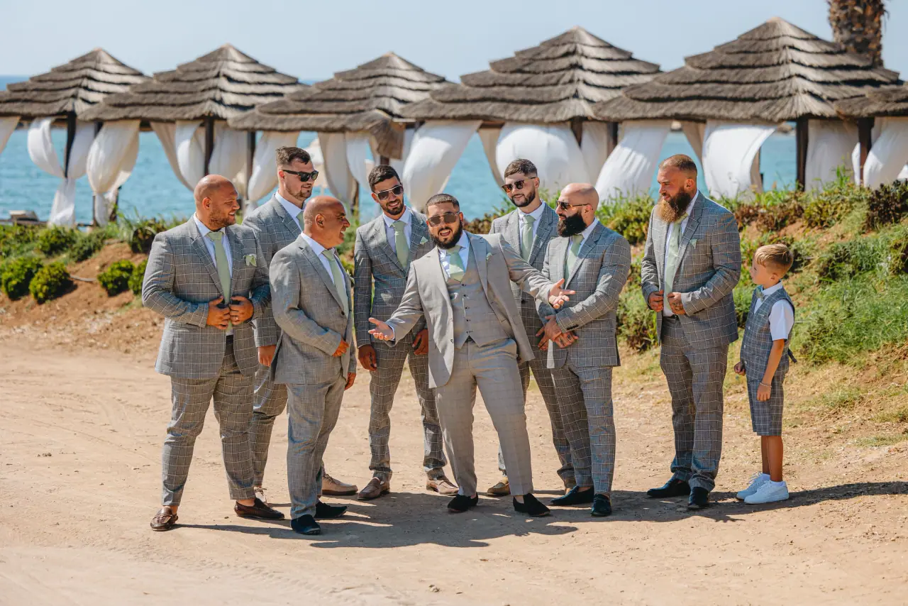 Groomsmen in grey suits with a page boy beside thatched beach parasols