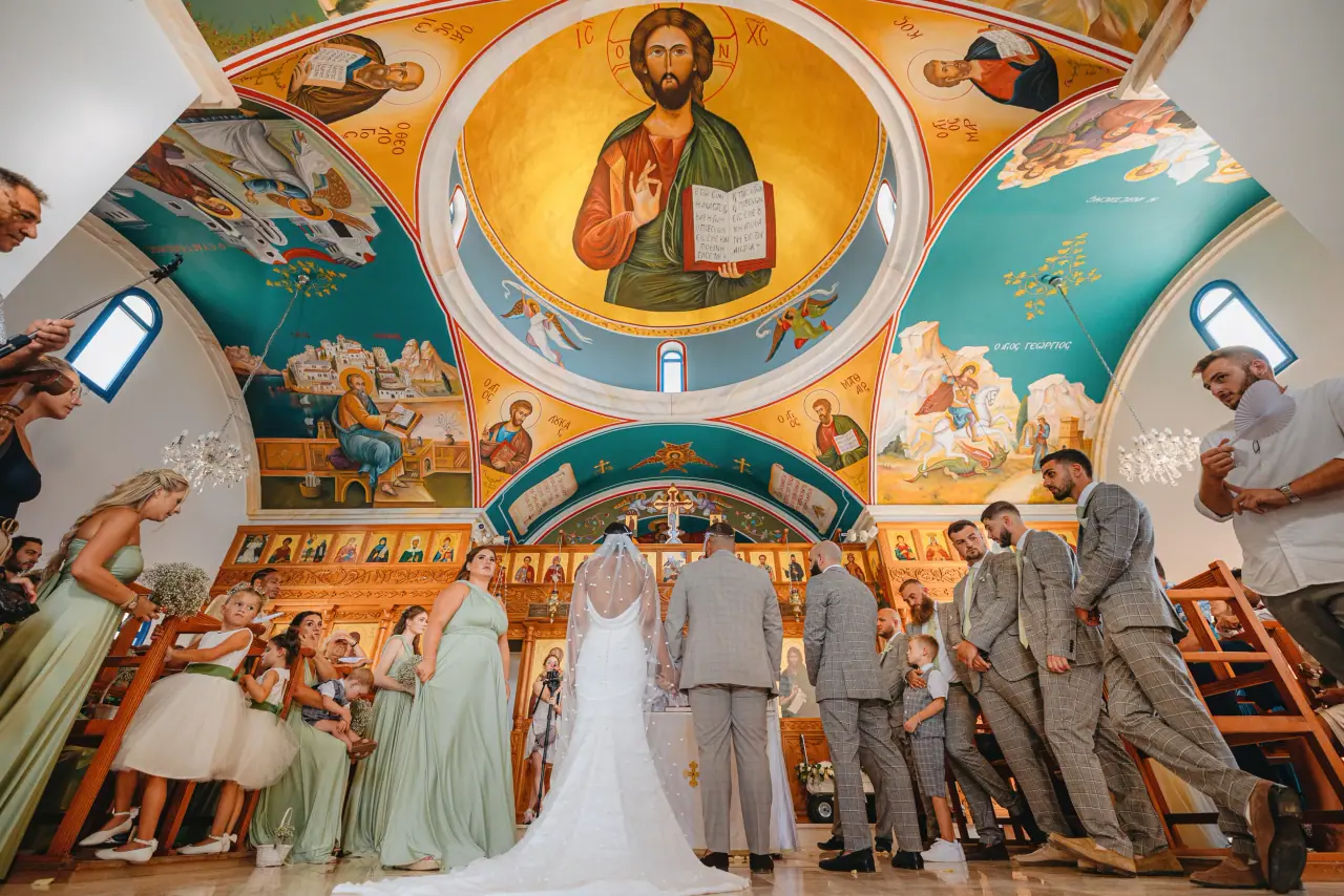 Wide view of the ceremony in a frescoed Orthodox church, bridesmaids in sage green