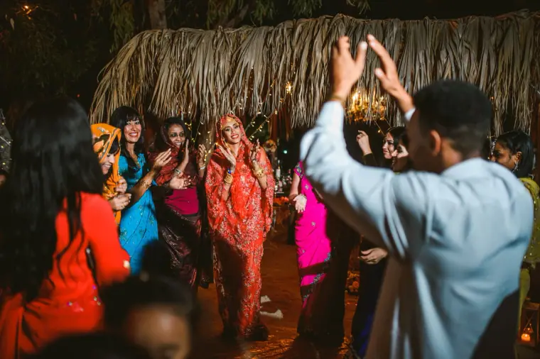 Guest raising his arms as the bride dances among women in bright outfits