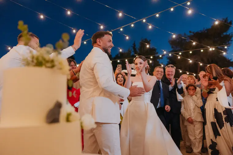 Bride and groom celebrating by the cake under festoon lights as guests cheer