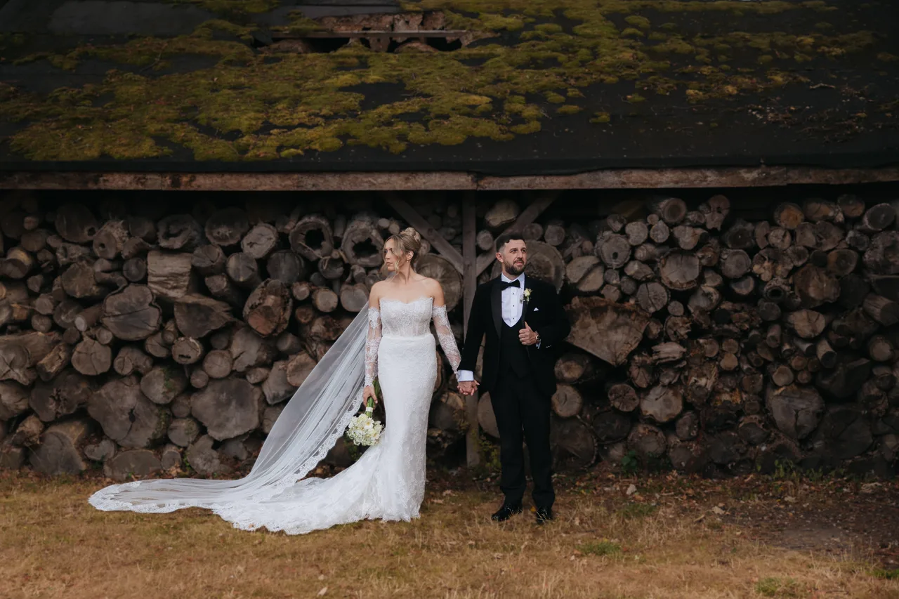 Bride and groom walking hand in hand past a moss-roofed woodshed at Northbrook Park, UK