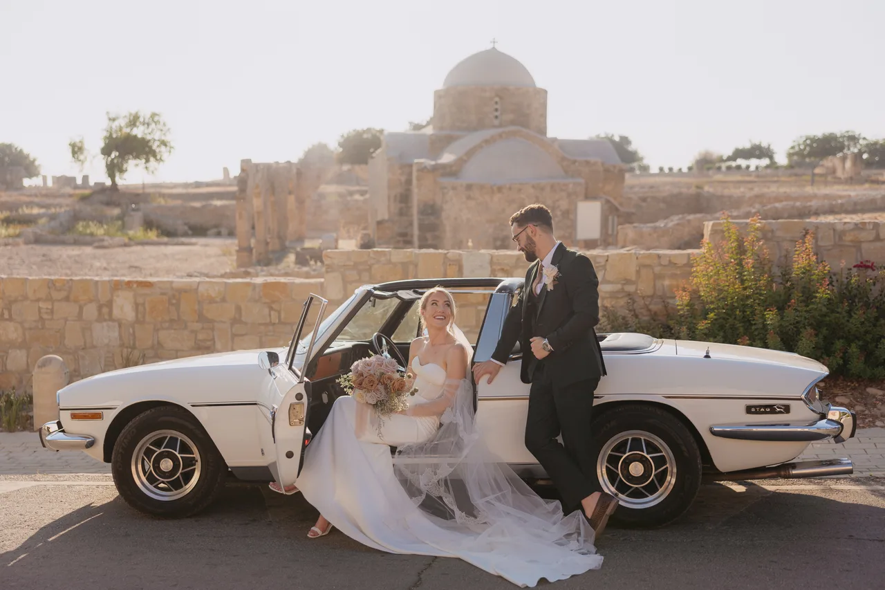 Bride seated in the 1977 Triumph Stag beside the groom, Agia Kyriaki church and ruins behind, Paphos
