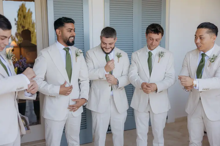 Groomsmen in cream suits and green ties laughing while getting ready