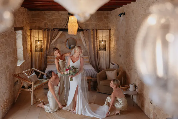 Bridesmaids helping the bride into her gown in a stone-walled suite at Liopetro, Kouklia