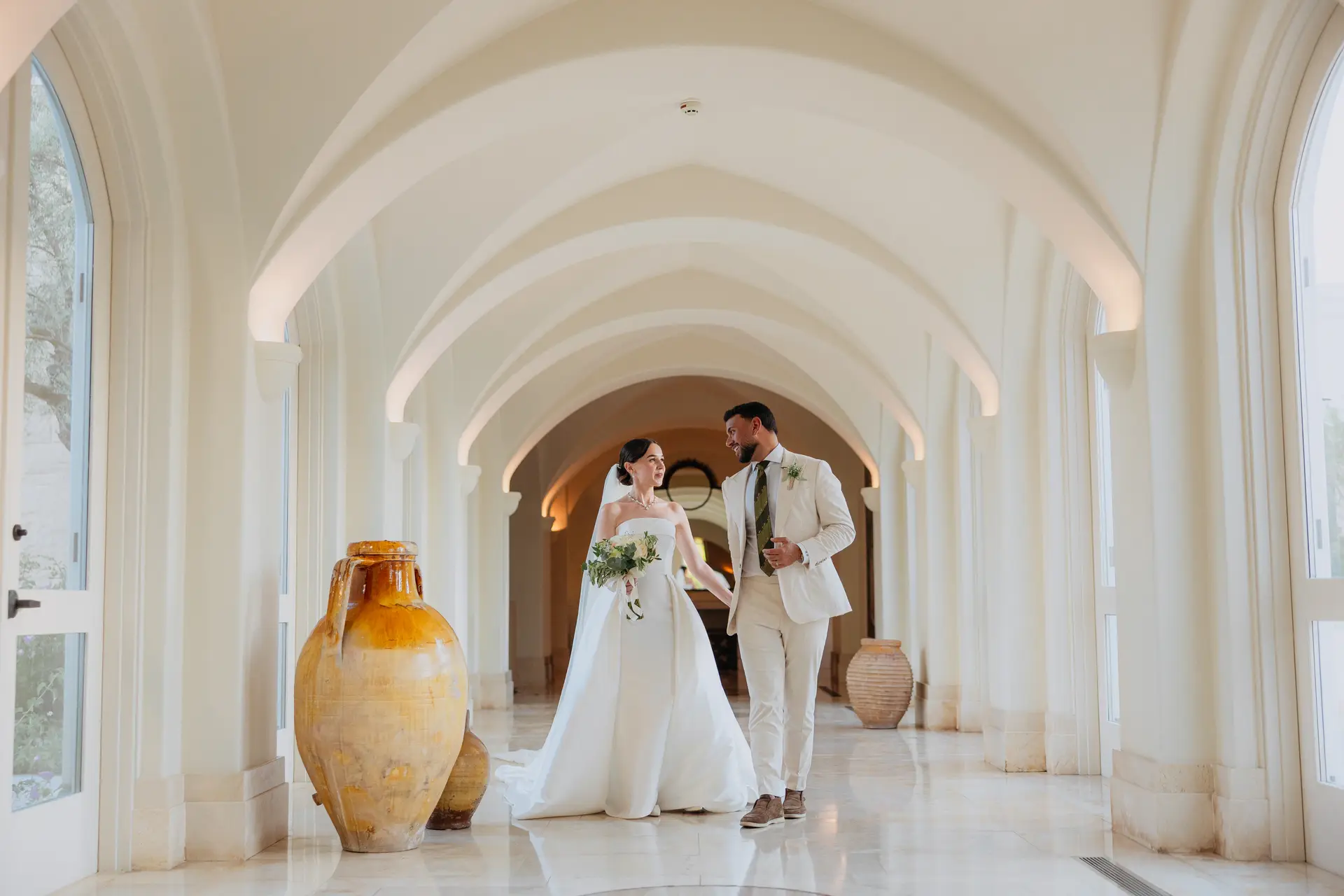 Poppy and Andrew walking hand in hand through Anassa's white vaulted cloister