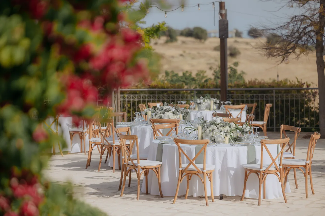 Round tables with white linen and cross-back chairs seen past red bougainvillea at Liopetro